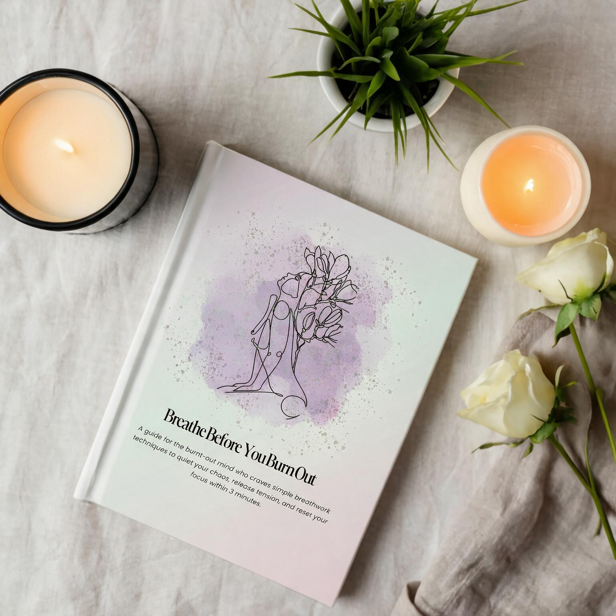 A book titled 'Breathe Before You Burn Out' lies on a light fabric surface, styled with a lit candle, a small potted plant, another candle, and white roses. The cover has a pale pink background with a lavender watercolor splash and a simple line drawing of a woman meditating