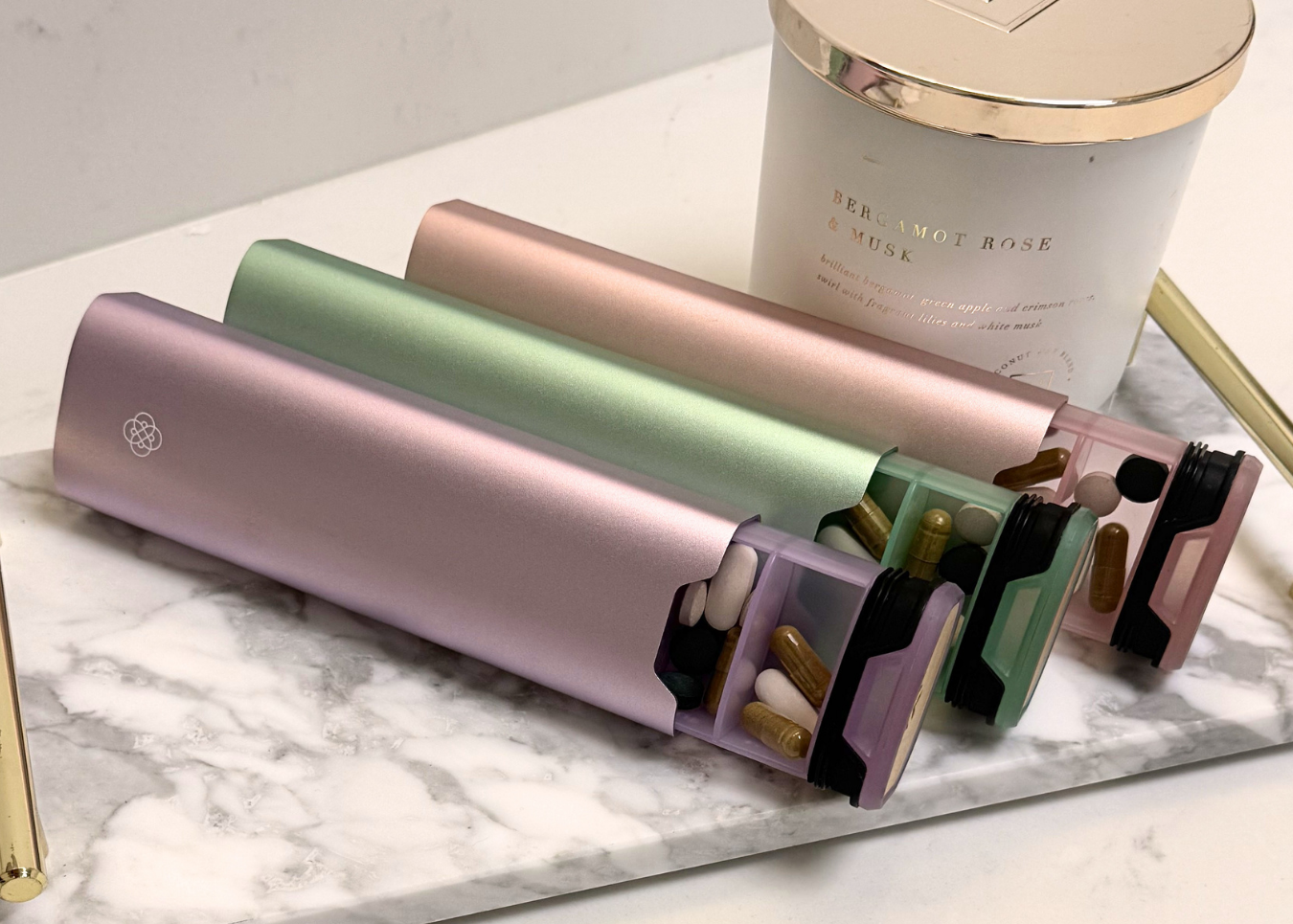 Three pillbox organisers (purple, green and pink) on a marble surface with a candle in the background.