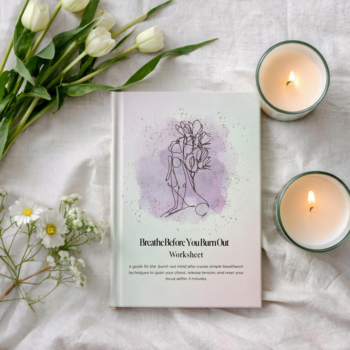 A book or workbook titled "Breathe Before You Burn Out Worksheet" lies on white fabric, surrounded by white tulips, small white flowers, and two lit candles in green-rimmed glass holders. The cover has a soft lavender watercolor splash with a simple line drawing of a woman meditating.